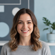 A smiling woman in her late 20s, with bright eyes, looking directly at the viewer. She wears a casual top. The background is 